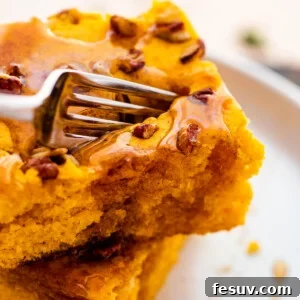 A fork cuts into a fluffy, golden-brown sheet pan pumpkin pancake, revealing its soft interior and inviting texture. A dusting of powdered sugar and a drizzle of maple syrup glisten on top.