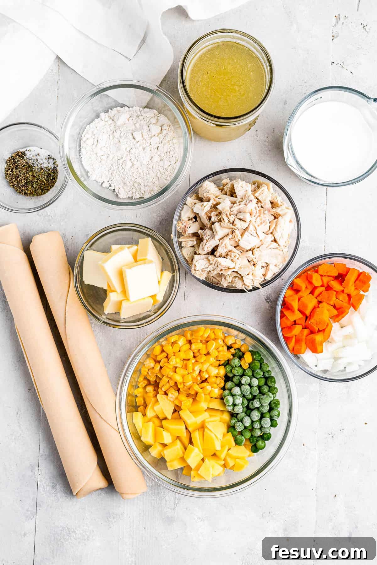 A selection of fresh ingredients for chicken pot pie, arranged clockwise: golden chicken broth, creamy milk, freshly chopped carrots and onions, a bowl of bright green peas, corn, and finely diced potatoes, a stick of butter, a variety of seasonings, and all-purpose flour.