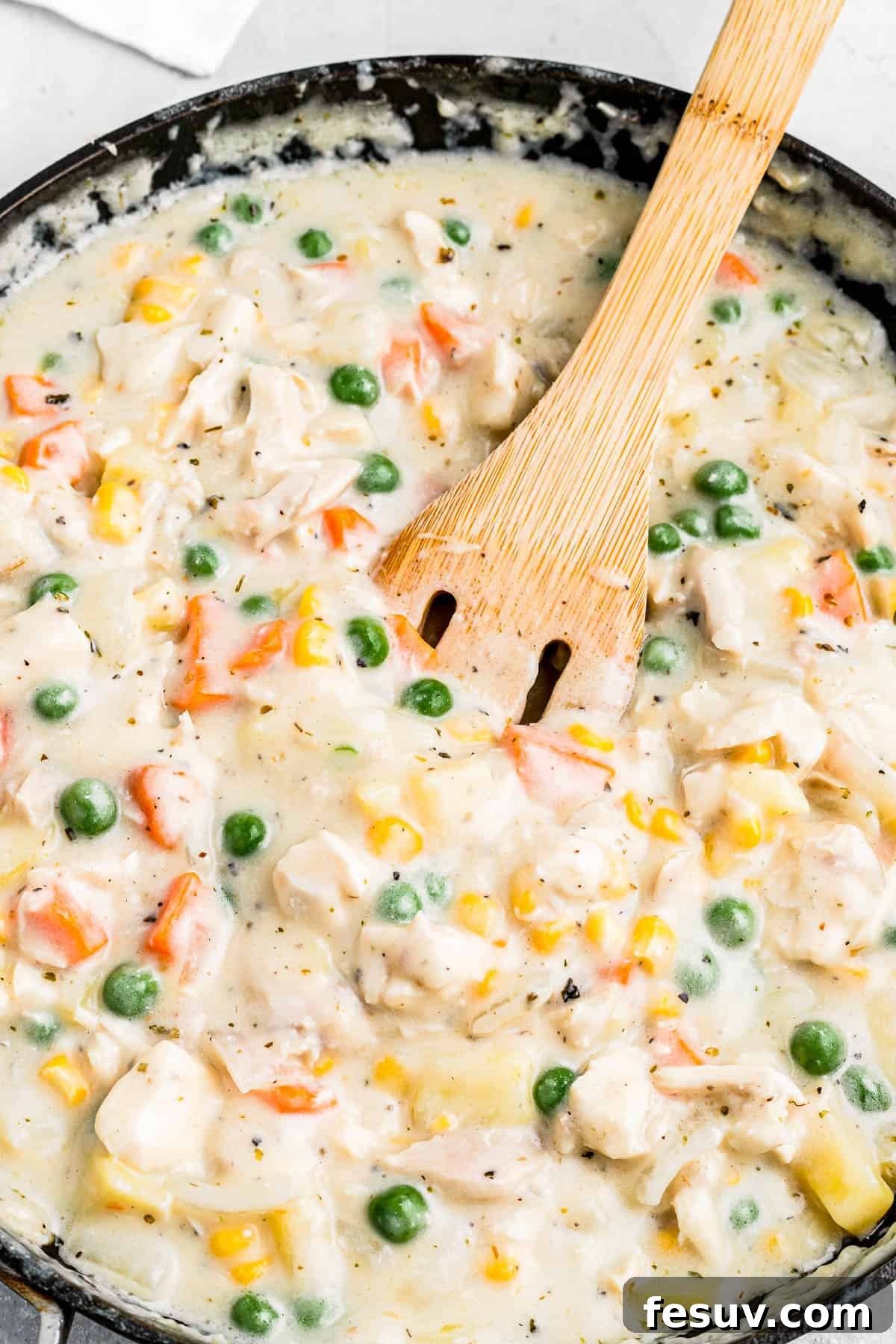 A wooden spoon stirring a creamy chicken pot pie filling in a skillet, showcasing the rich gravy, tender chicken pieces, bright carrots, green peas, golden corn, and soft diced potatoes, all simmering together.