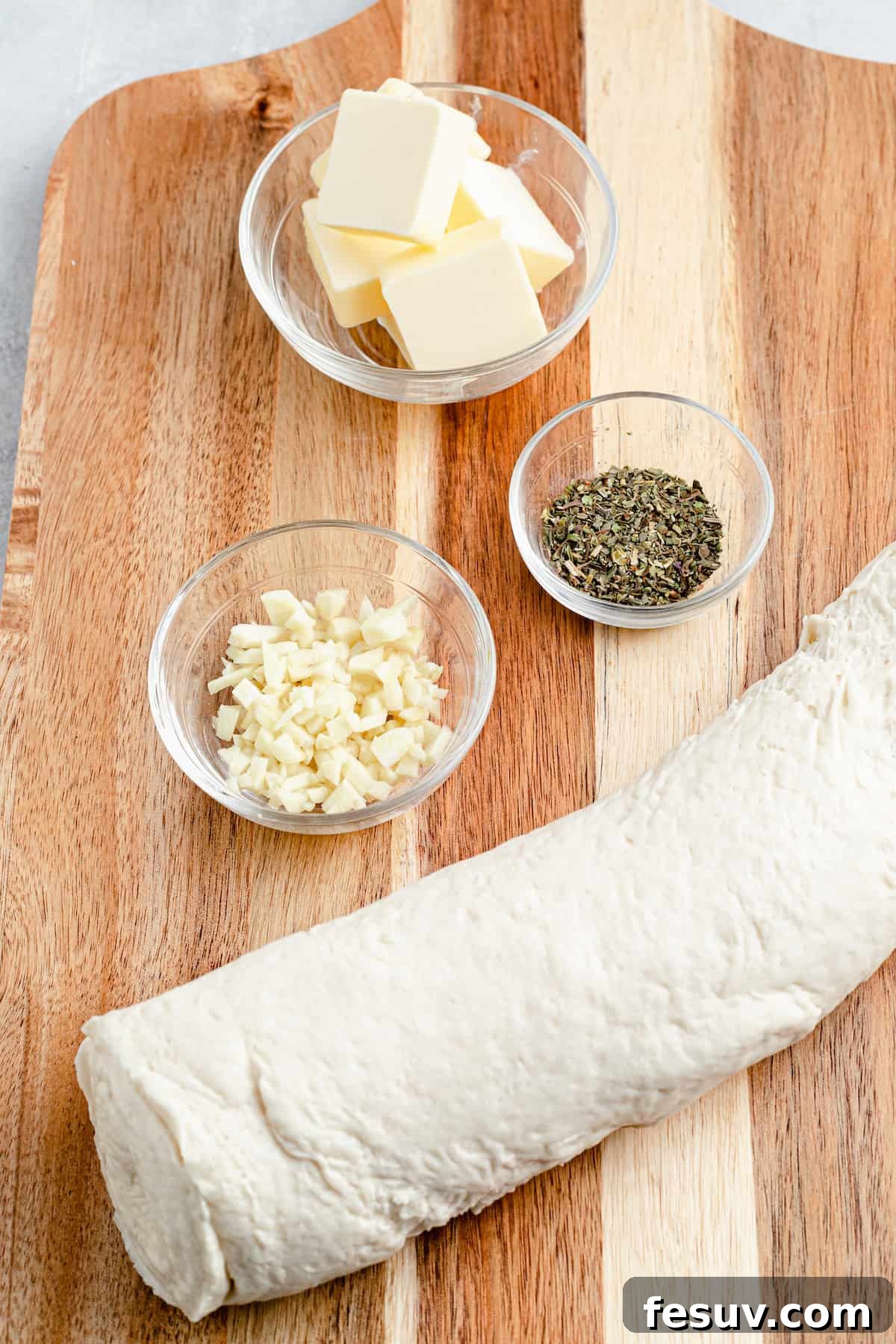 Key ingredients for homemade garlic knots: a stick of butter, a shaker of Italian seasoning, a roll of refrigerated pizza dough, and a small dish of freshly minced garlic.