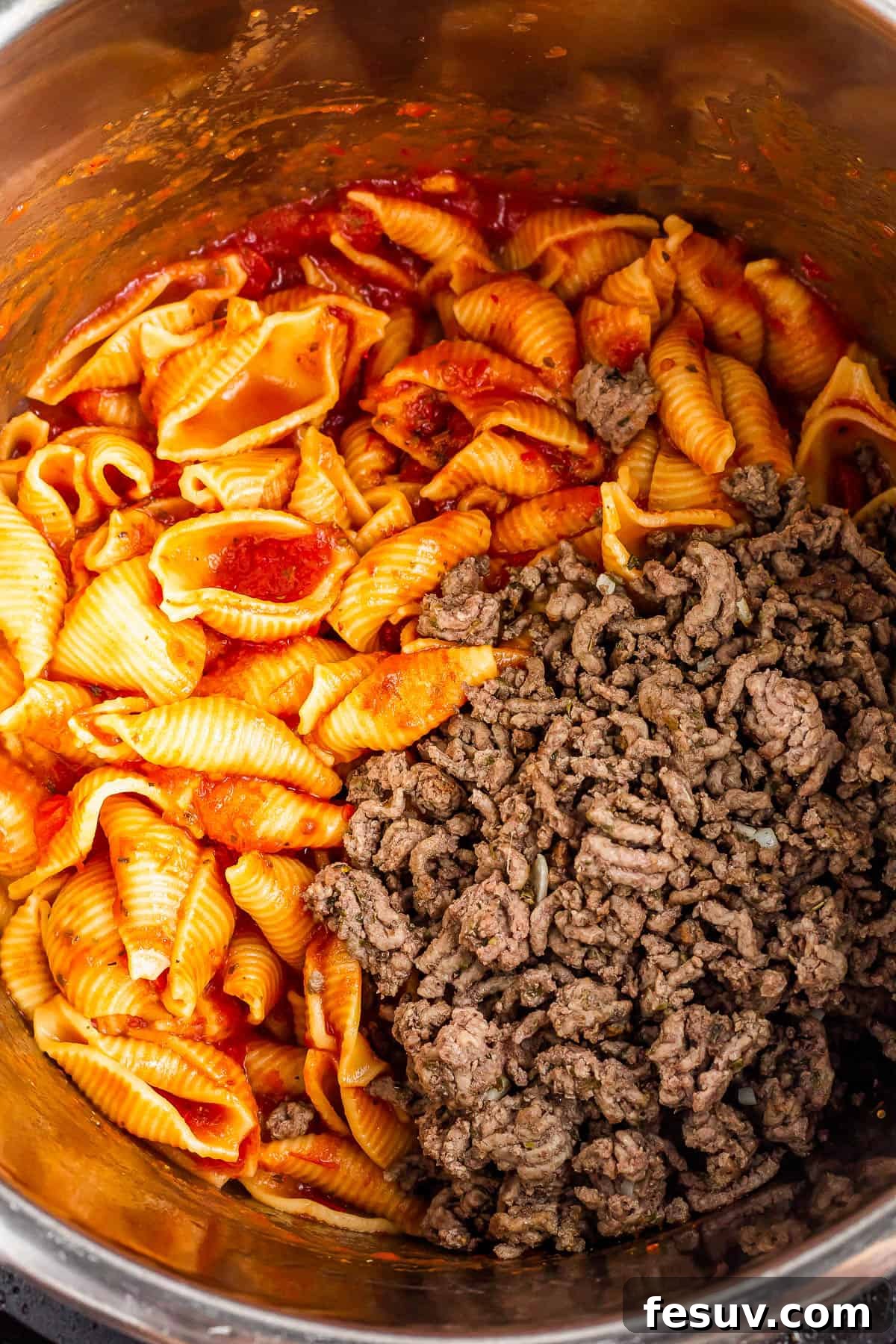 An overhead shot of an Instant Pot with cooked shells in sauce inside and a batch of browned ground beef added, ready to be stirred.