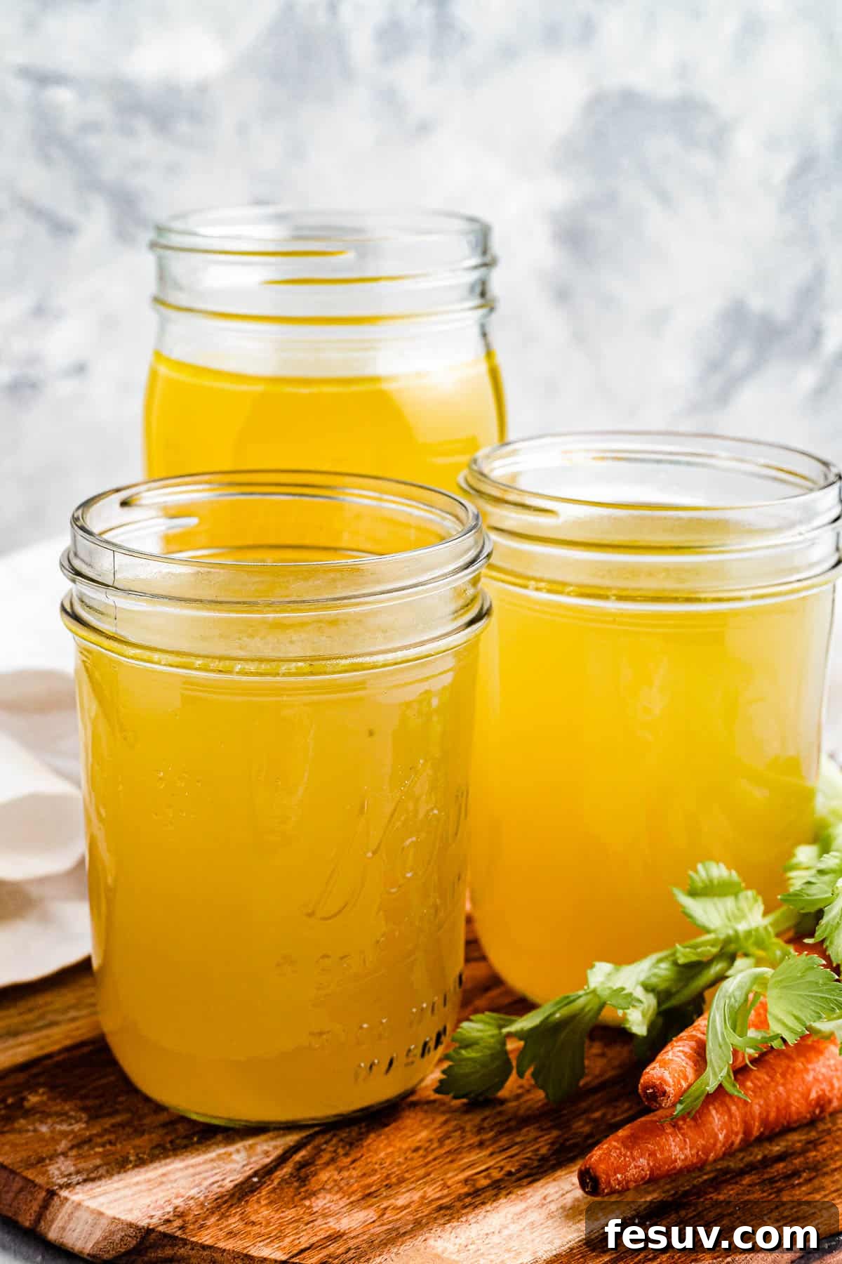 Homemade Instant Pot Chicken Broth 2 Three jars of homemade chicken broth on a cutting board with raw carrots nearby, symbolizing freshness and homemade goodness.