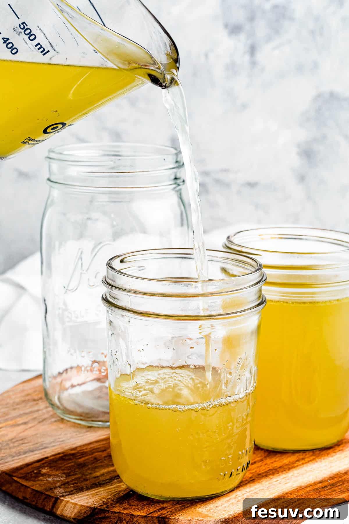 Homemade Instant Pot Chicken Broth 3 Chicken broth being poured from a large measuring cup into several jars, highlighting the process of preserving homemade broth.