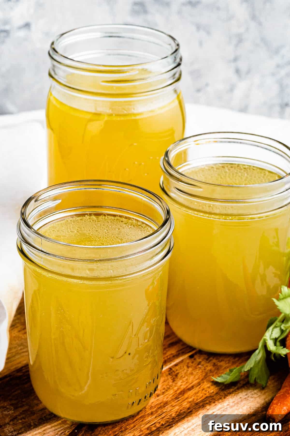 Homemade Instant Pot Chicken Broth 7 Three jars of chicken broth on a cutting board with raw carrots positioned next to them, emphasizing the fresh, homemade quality.