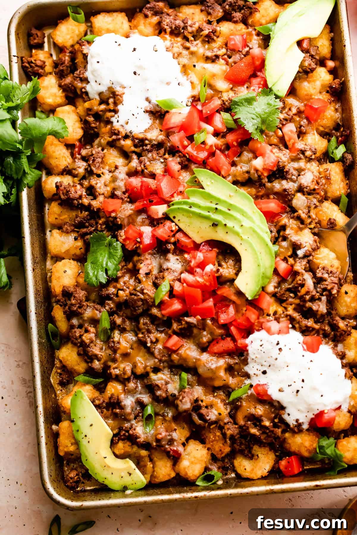 Loaded Sheet Pan Beef Totchos 2 Crispy tater tots topped with seasoned ground beef, melted cheese, fresh diced tomatoes, a dollop of sour cream, and sliced avocado on a sheet pan, ready to serve.