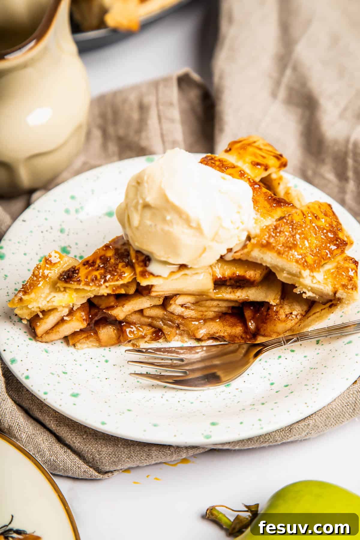 This classic yet easy and delicious Apple Pie recipe is made from scratch, with a buttery, flaky pie crust, fresh apple slices, pie spices, and brown sugar. A perfectly baked slice of homemade apple pie, topped with a scoop of melting vanilla ice cream, served on a white plate with a fork beside it. The crust is golden brown and flaky.