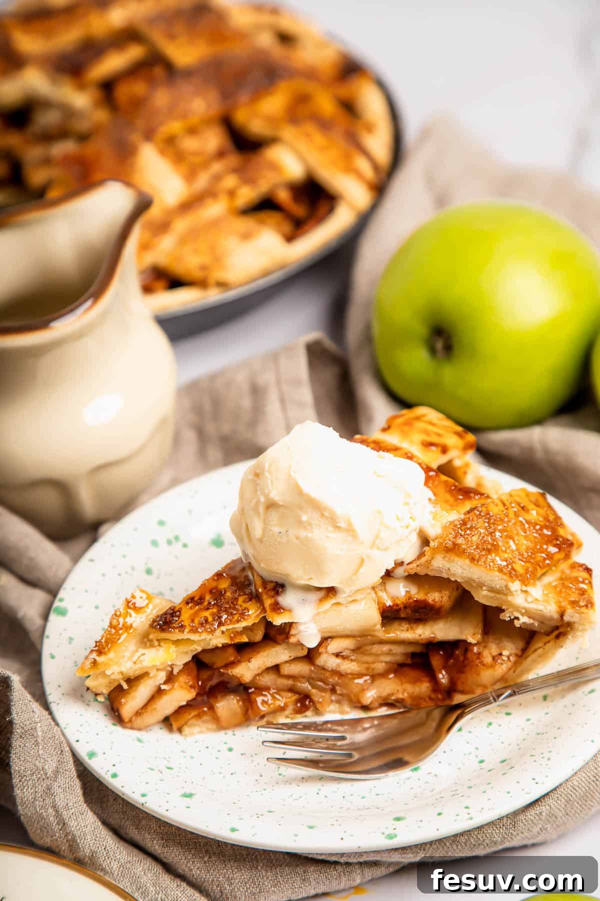 This classic yet easy and delicious Apple Pie recipe is made from scratch, with a buttery, flaky pie crust, fresh apple slices, pie spices, and brown sugar. A close-up shot of a single slice of warm apple pie, generously topped with a scoop of vanilla ice cream, on a white plate with a silver fork beside it.