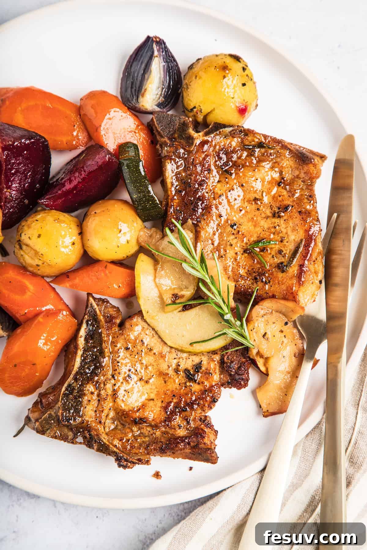 A beautifully plated baked pork chop with roasted apples and assorted winter vegetables, garnished with a fresh sprig of rosemary.