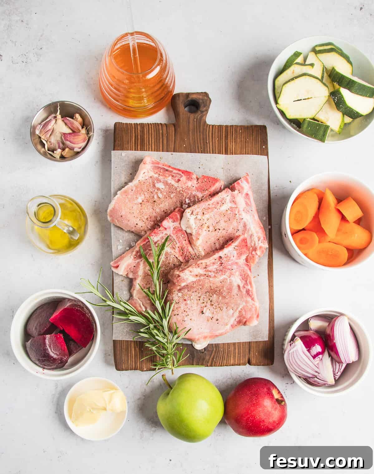 An array of fresh ingredients for baked pork chops: sliced zucchini, chopped carrots, chopped onions, two whole apples, butter, beetroot, oil, garlic cloves, apple cider, four seasoned pork chops, and a sprig of fresh rosemary, arranged neatly on a wooden surface.