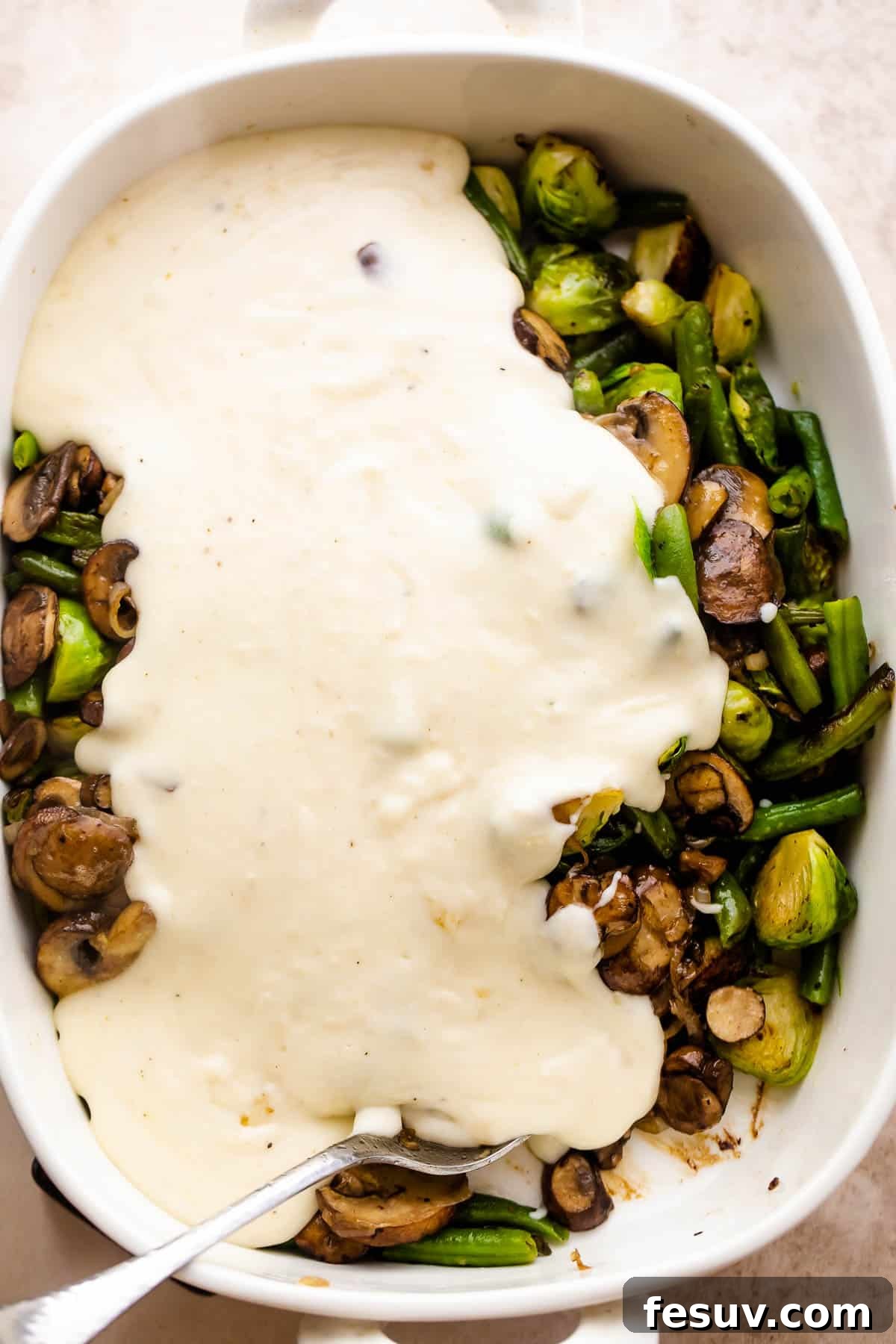 Assembling the Green Bean Casserole A rich, creamy Parmesan sauce being poured over a bed of sautéed green beans, Brussels sprouts, and mushrooms in a white baking dish, ready for the oven.