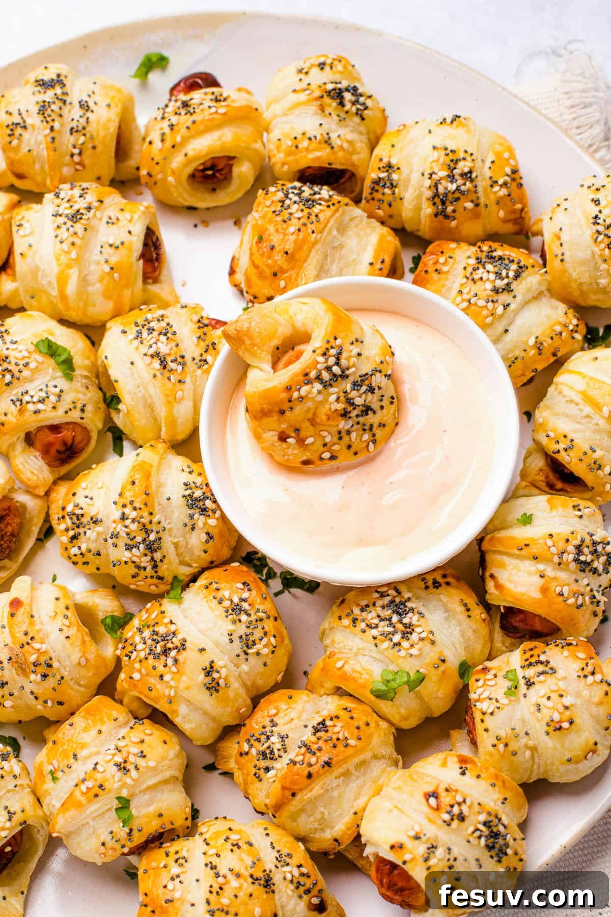 Super easy to make homemade Pigs in a Blanket are a delicious snack prepared with savory mini-sausages wrapped in golden, flaky puff pastry. A beautifully arranged platter of golden-baked pigs in a blanket, with a small bowl of a creamy, reddish dipping sauce at the center. One pastry is already dipped, showing its delicious coating.