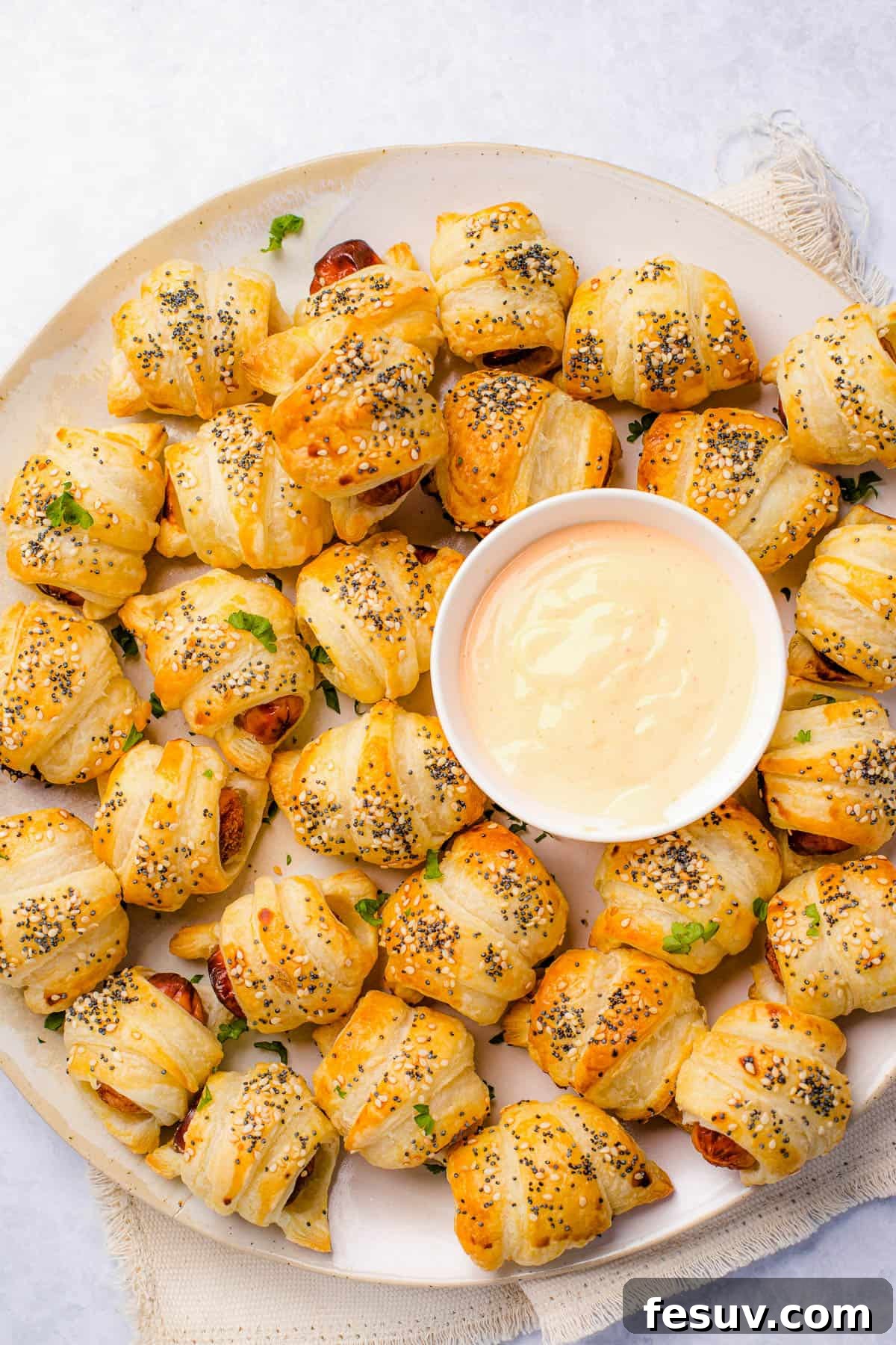 Super easy to make homemade Pigs in a Blanket are a delicious snack prepared with savory mini-sausages wrapped in golden, flaky puff pastry. A vibrant close-up of a platter showcasing freshly baked pigs in a blanket, golden and sprinkled with seeds, artfully arranged around a small white bowl filled with a creamy, reddish dipping sauce, ready to be enjoyed.