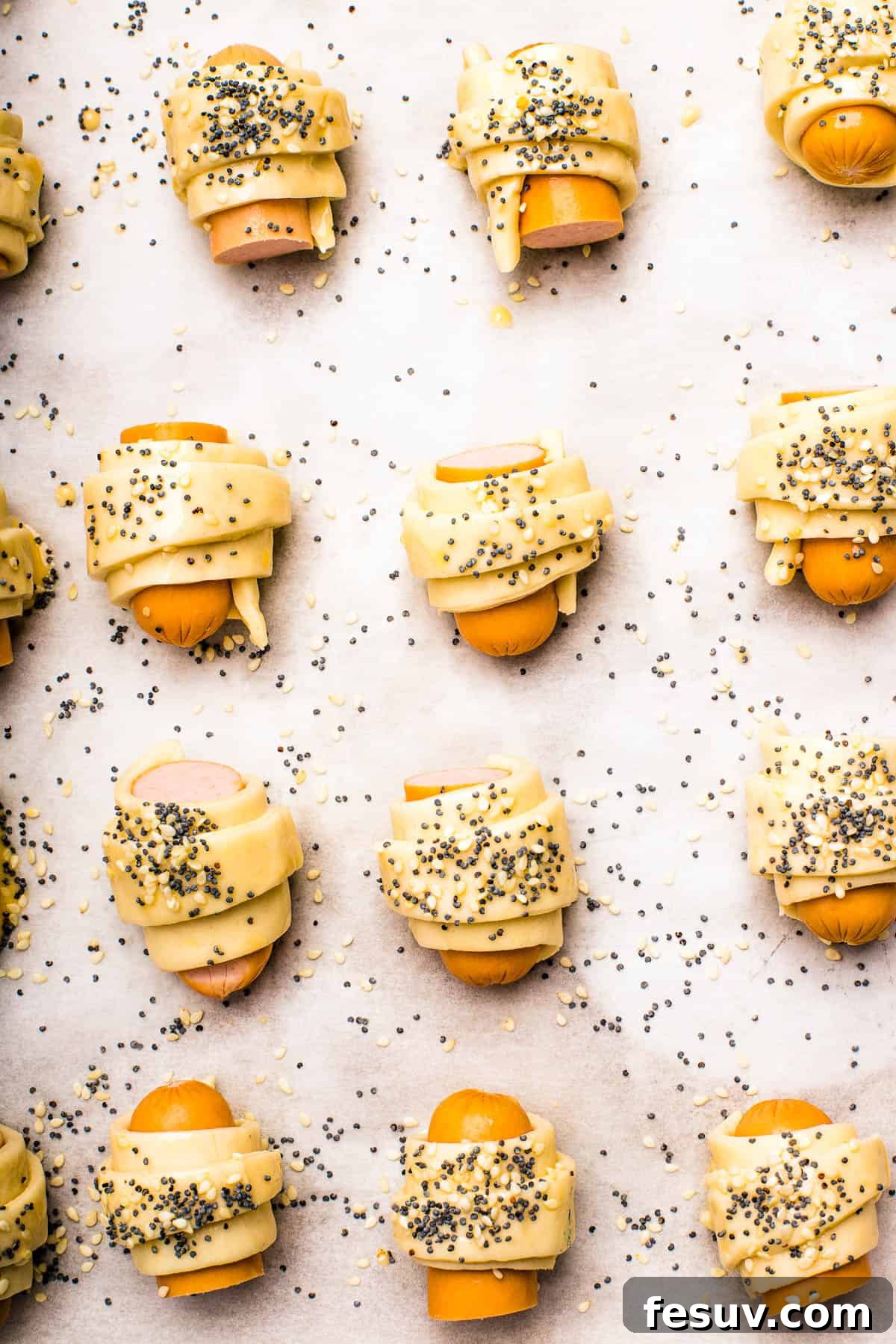 Super easy to make homemade Pigs in a Blanket are a delicious snack prepared with savory mini-sausages wrapped in golden, flaky puff pastry. Close-up shot of unbaked pigs in a blanket, neatly arranged on parchment paper, each brushed with egg wash and generously sprinkled with a mix of sesame and poppy seeds, ready for the oven.