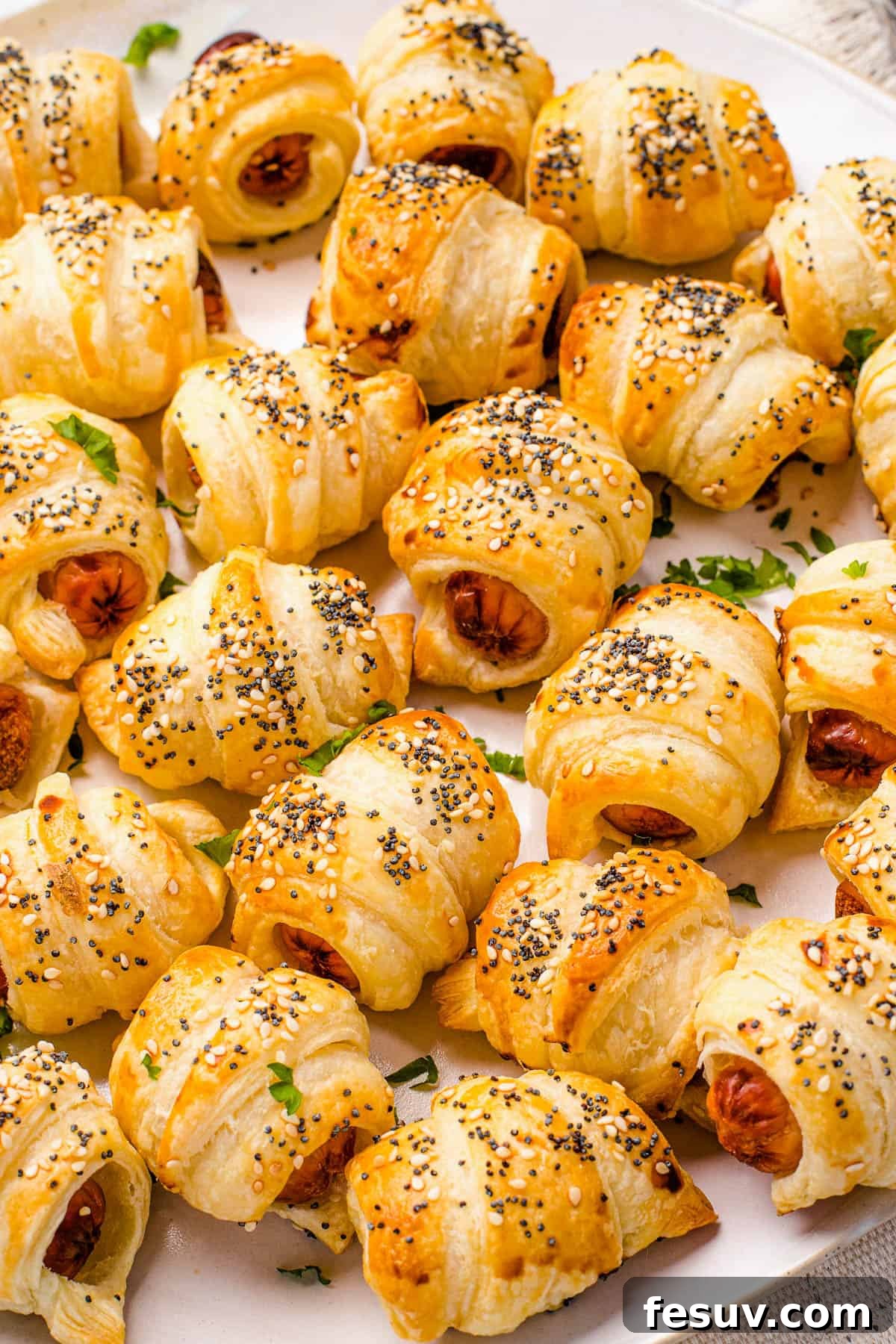 Super easy to make homemade Pigs in a Blanket are a delicious snack prepared with savory mini-sausages wrapped in golden, flaky puff pastry. A close-up view of crescent roll hot dogs, perfectly golden brown after baking, showcasing the flaky pastry texture and the appealing sprinkle of sesame and poppy seeds on top.