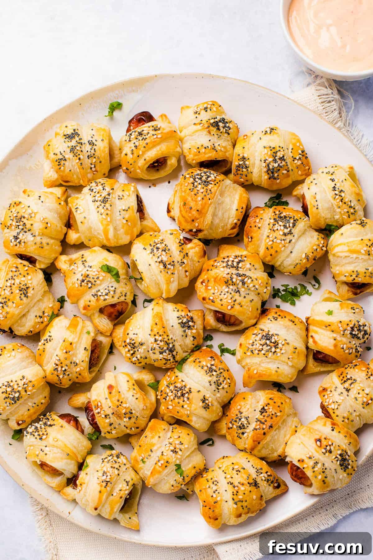 Super easy to make homemade Pigs in a Blanket are a delicious snack prepared with savory mini-sausages wrapped in golden, flaky puff pastry. A festive serving platter showcasing an abundant arrangement of perfectly baked, golden pigs in a blanket, ready to be enjoyed as appetizers. A small bowl of dipping sauce is placed conveniently nearby.