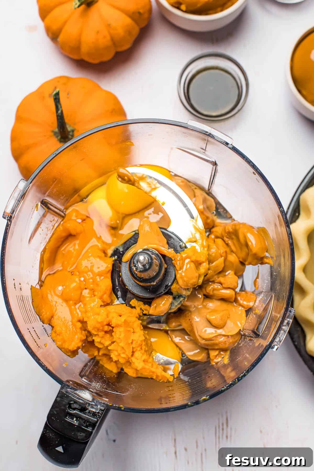 All the vibrant pumpkin pie filling ingredients, including pumpkin puree and spices, gathered in a blender on a white countertop, surrounded by small decorative pumpkins.