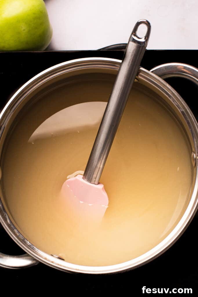 A saucepan of undyed sugar candy mixture with a spatula resting in it, showing the syrup's clear consistency before coloring.