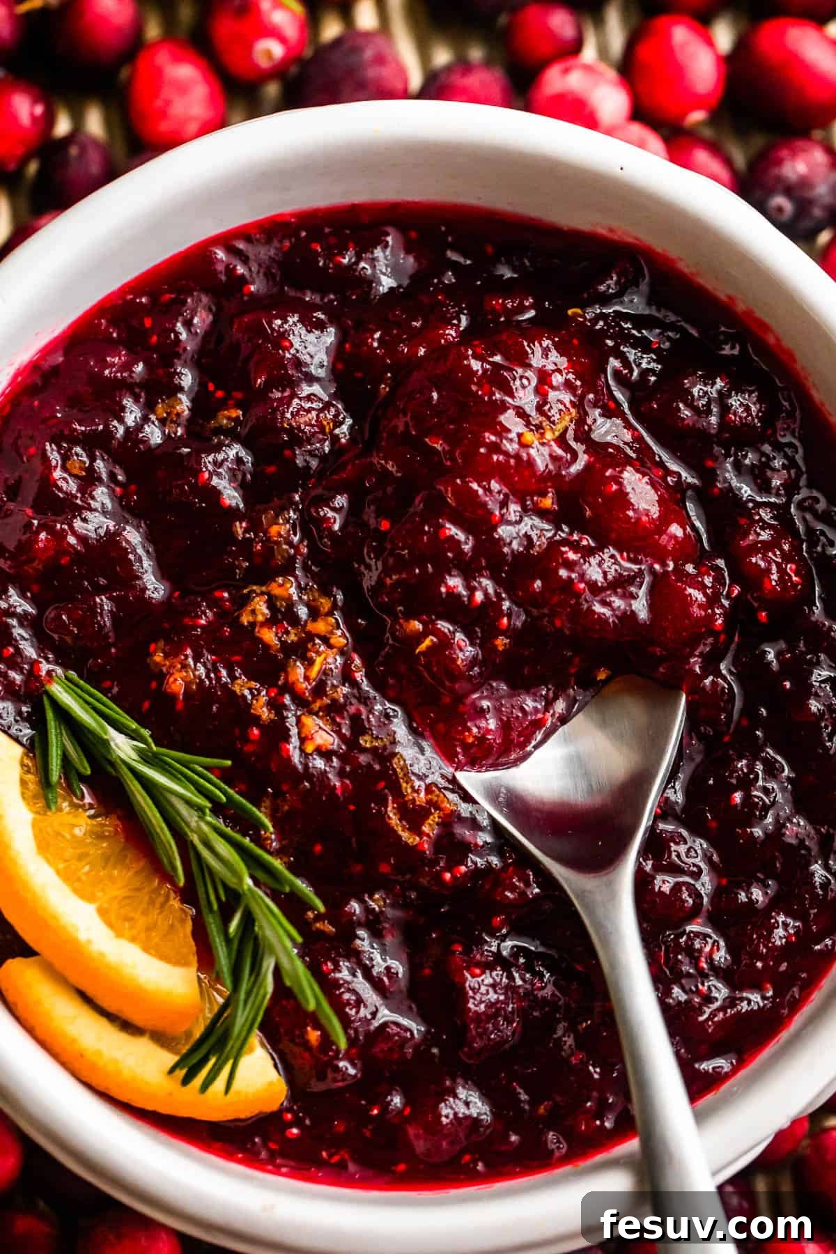 Divine Homemade Cranberry Sauce 2 overhead close up shot of a spoon inside of a bowl filled with cranberry sauce and garnished with rosemary sprig plus orange slices