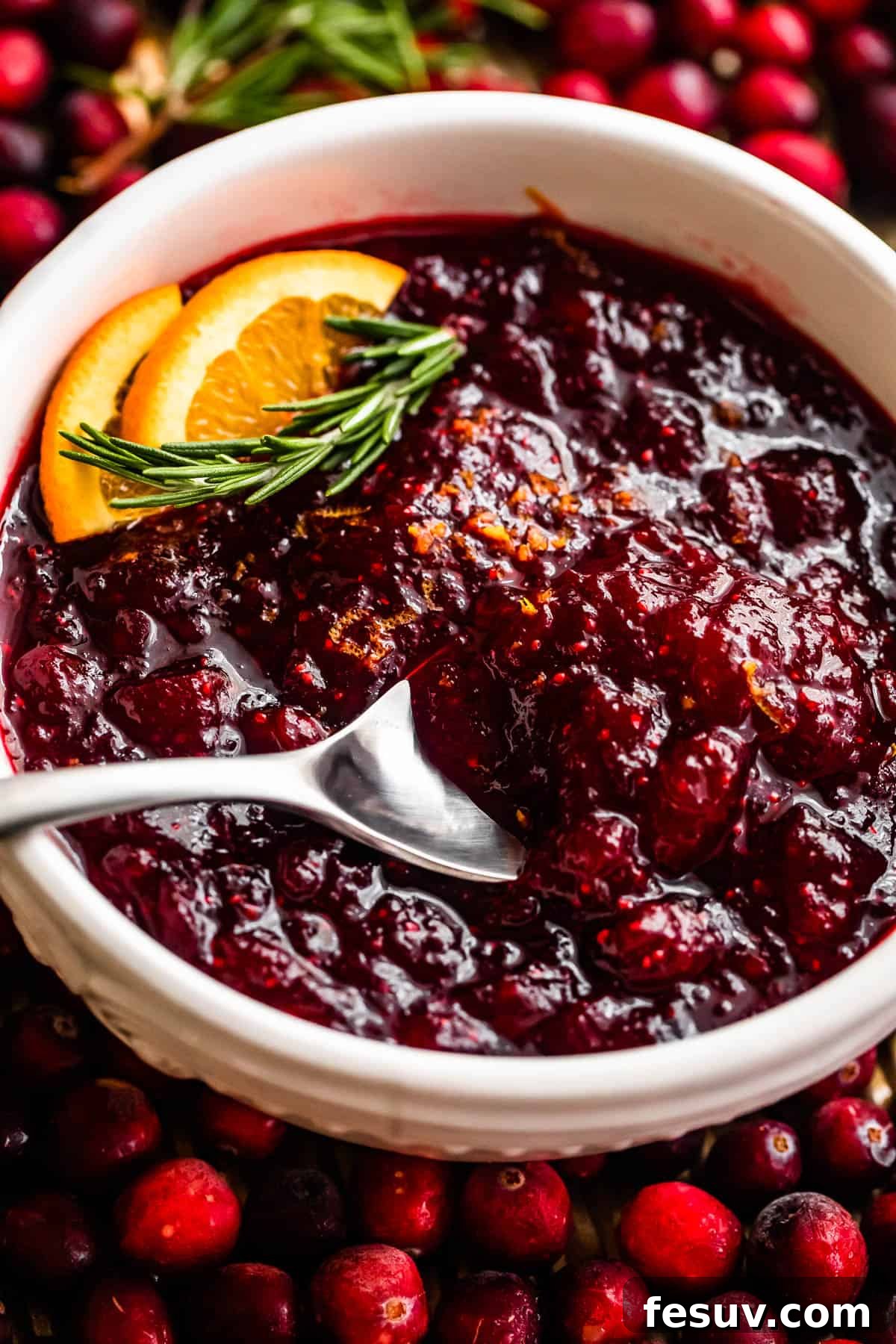 Divine Homemade Cranberry Sauce 3 cranberry sauce inside of a white bowl surround by fresh cranberries and garnished with orange slices and rosemary sprig.