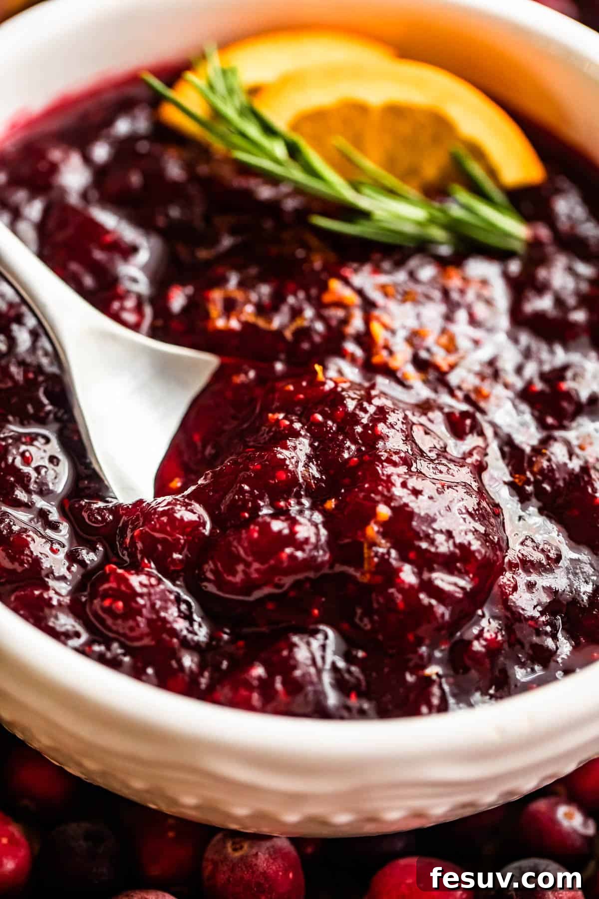 Divine Homemade Cranberry Sauce 6 side shot of a spoon inside of a bowl filled with cranberry sauce and garnished with rosemary sprig plus orange slices