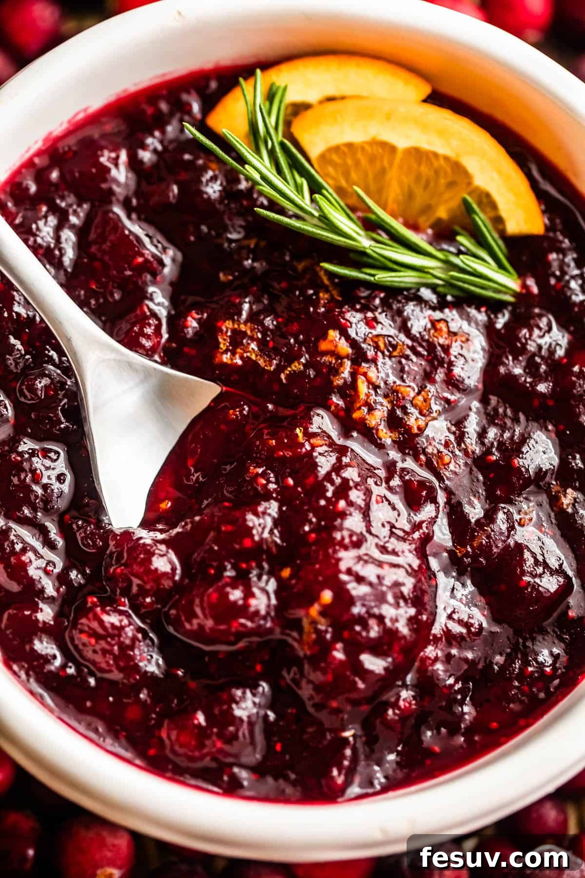 Divine Homemade Cranberry Sauce 7 overhead close up shot of a spoon inside of a bowl filled with cranberry sauce and garnished with rosemary sprig plus orange slices