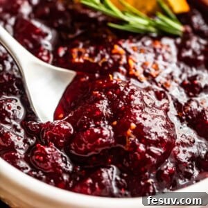 Divine Homemade Cranberry Sauce 8 side shot of a spoon inside of a bowl filled with cranberry sauce and garnished with rosemary sprig plus orange slices